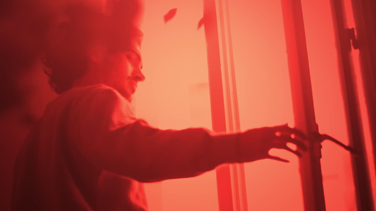 Hombre abriendo una ventana bajo una luz roja, comprobando el peligro exterior.