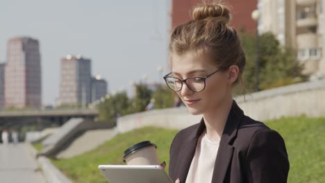 Geschäftsfrau-Mit-Brille-Und-Anzugjacke-Trinkt-Kaffee-Und-Schaut-Auf-Das-Tablet-Auf-Der-Straße