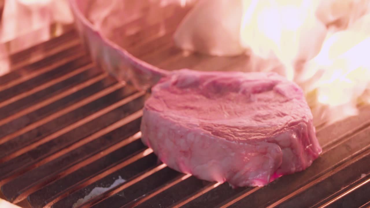 A slow-motion close-up shot highlights a juicy steak grilling over intense flames with onion slices, capturing the sizzling and smoky cooking process.