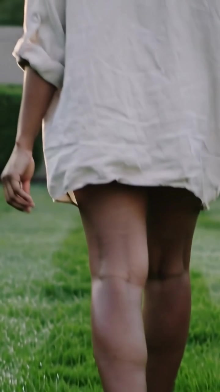 Video verticale: Una donna in camicia chiara inizia a camminare a piedi nudi sul prato, cercando la luce del sole.