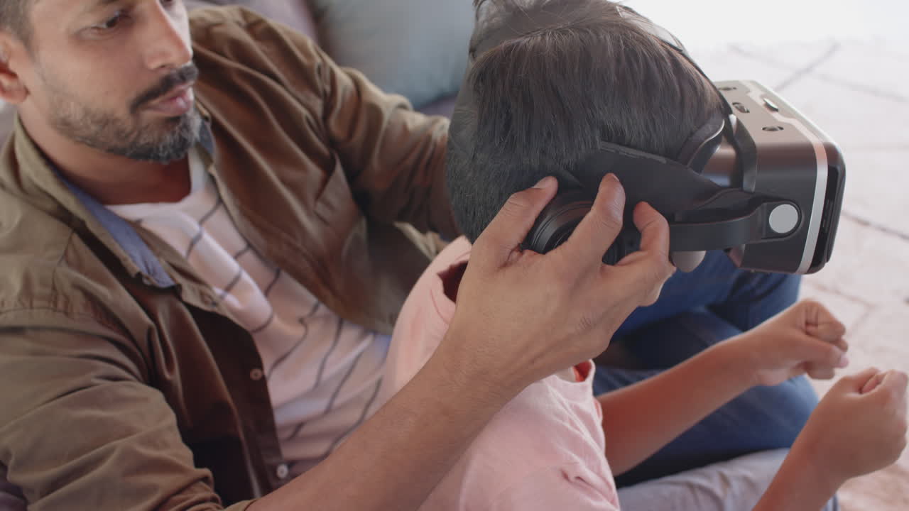 Using VR headset, young boy sitting with Indian father on couch, exploring virtual world
