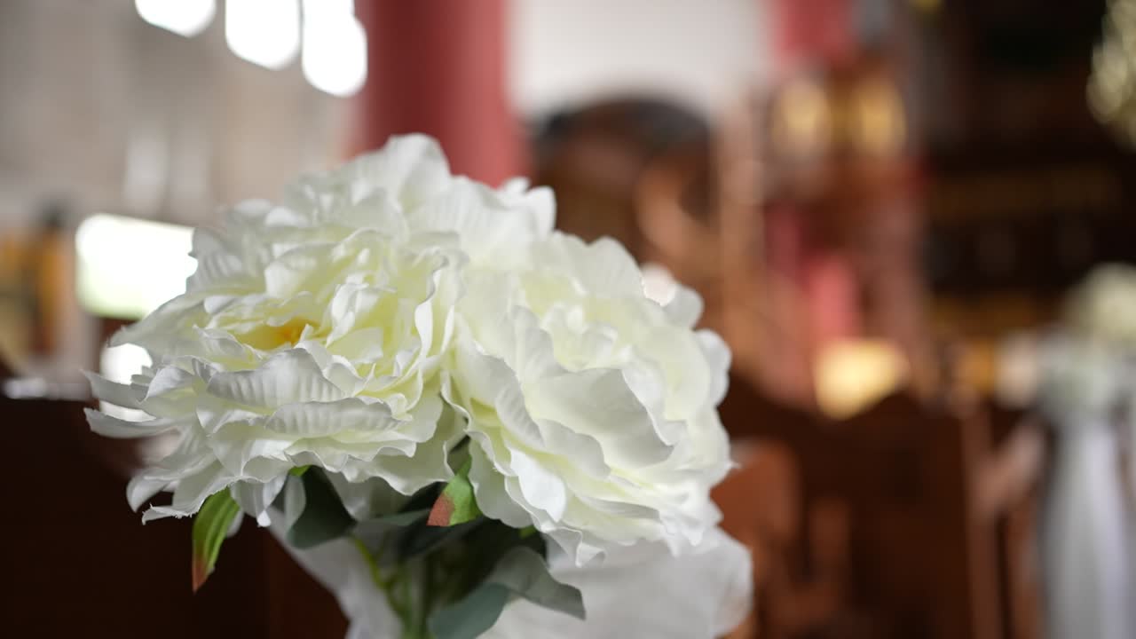Una veduta ravvicinata di fiori bianchi posati su un tavolo di legno in una chiesa. Le persone si riuniscono per una cerimonia. La luce filtra attraverso le finestre creando un'atmosfera calda.