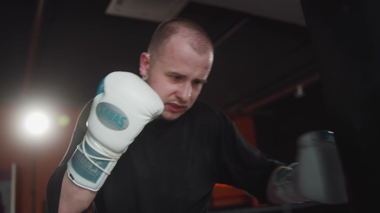 Bokser inde i et indendørs fitnesscenter leverer gentagne slag til en tung lædersæk, hvide polstrede handsker og sammenbidt kæbe viser anstrengelse, fokus, disciplin, styrke, varm belysning og dynamisk sløring omkring træningen