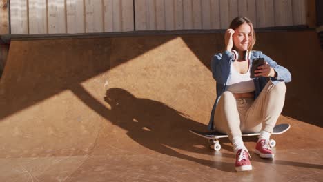 video of happy caucasian female skateboarder resting and using smartphone