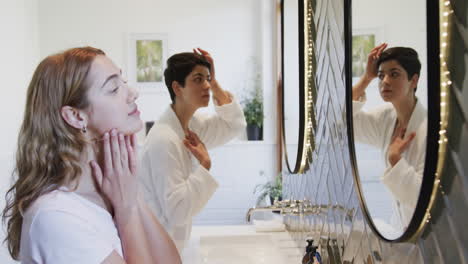 Happy-caucasian-lesbian-couple-looking-in-mirror,-improving-hair-and-smiling-in-bathroom