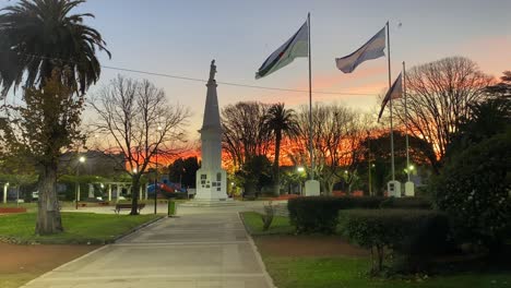 park with a white monument surrounded by trees, gardens, and waving flags, under a sky painted with the colors of sunset