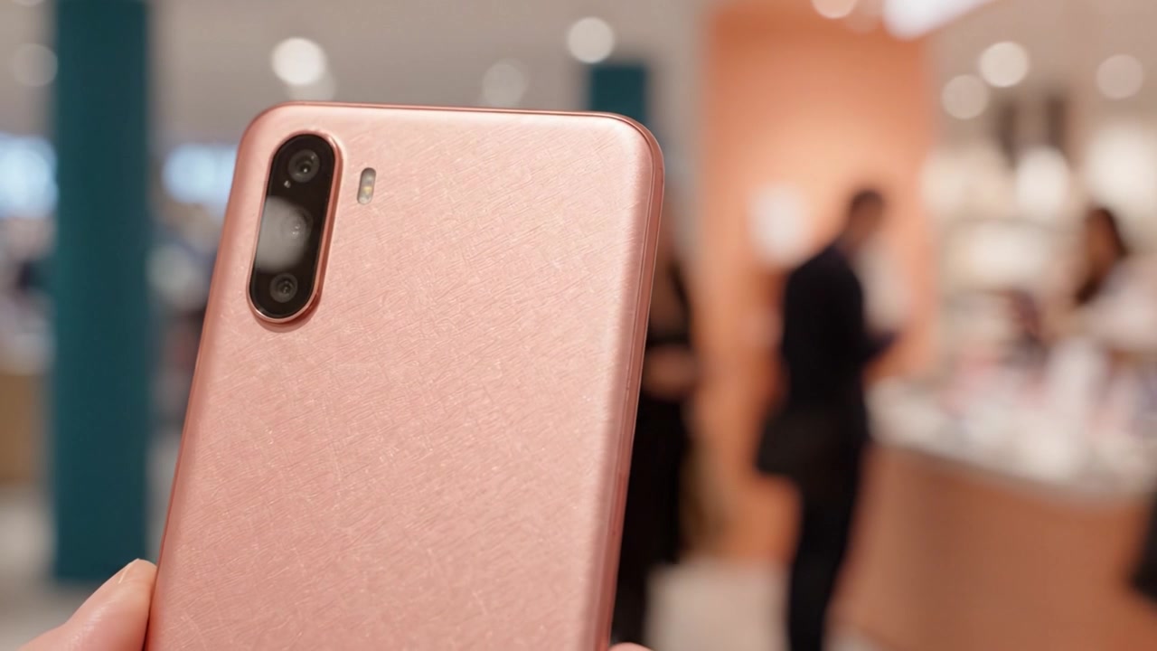 Detail moderního smartphonu s růžovými texturovanými zády, předvádějící jeho nastavení fotoaparátu, na rozmazaném pozadí maloobchodního prostředí plného lidí