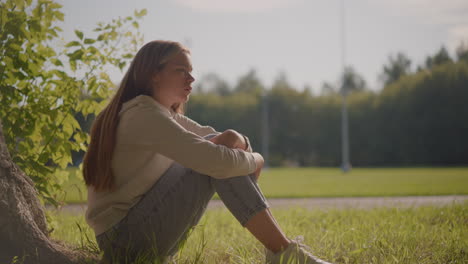 close-up side view of woman seated on grassy field, deeply lost in thought, with sunlight illuminating her face and arms folded over her knees, blurred stadium poles and greenery in the background