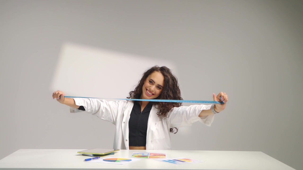 Retrato de medio cuerpo de una nutricionista en su escritorio estirando una cinta métrica y sonriendo ampliamente sobre fondo blanco. Concepto de medición corporal, orientación sobre estilo de vida saludable y consulta segura.