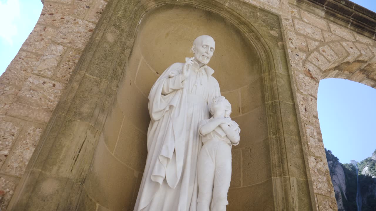 A white stone sculpture of a saint guiding a child, situated in an arched stone niche at the Montserrat monastery. This religious artwork is part of the monastery's extensive collection of outdoor sta