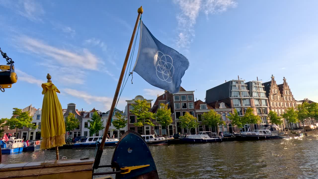 Blue flag flutters on boat, canal view, historic Haarlem architecture, sunny summer daylight, steady shot