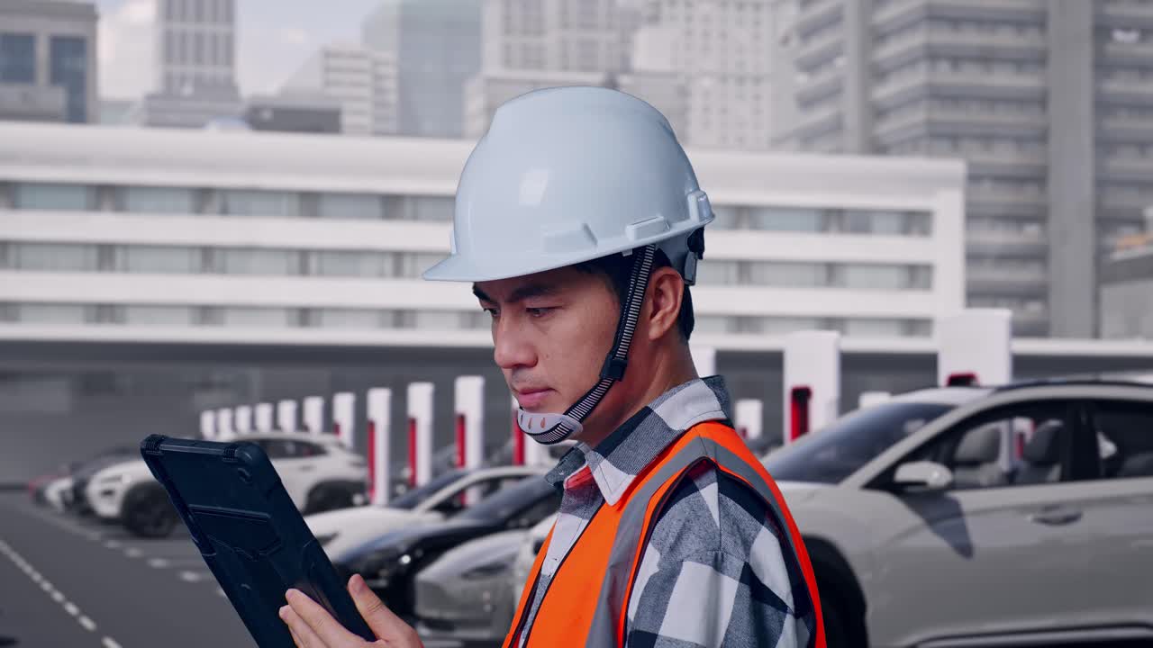 Primo piano laterale di un ingegnere uomo asiatico con casco di sicurezza che lavora su un tablet mentre è in piedi, con veicoli elettrici in ricarica presso una moderna stazione di ricarica urbana