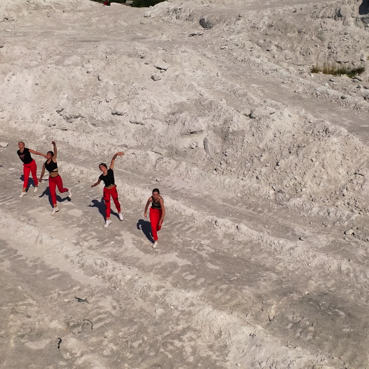 Aktif bir şekilde birlikte hareket eden, yan yana sıralanmış altı kız. Dans stüdyosu, kayalık doğa manzarası içinde performans sergiliyor. Üstten görünüm