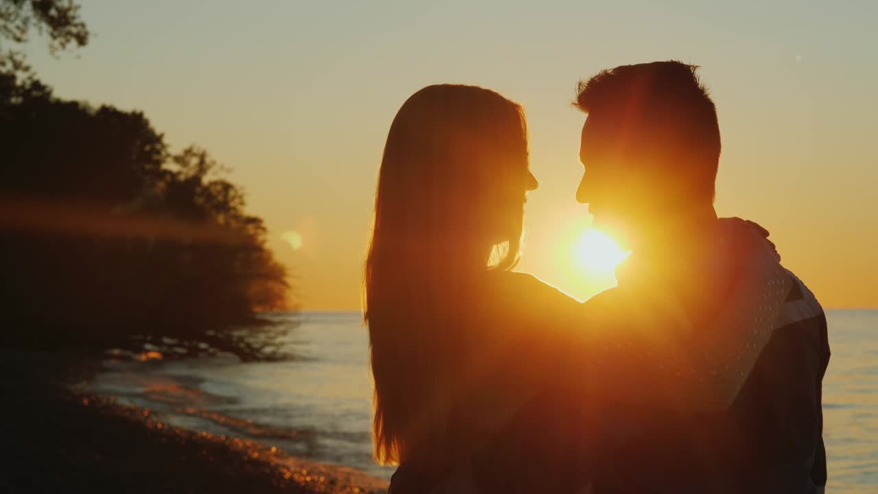 una pareja romántica multiétnica abrazándose al atardecer, de pie frente al mar, mirándose el uno al otro.