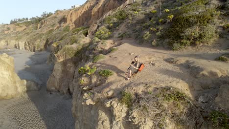 Woman-Sitting-in-Chair-by-Sea-Cliff-Reading-a-Book-in-Peace---Aerial-Orbit