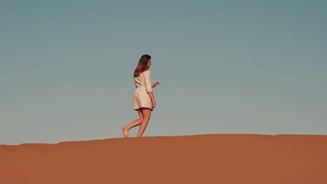 Young-caucasian-woman-walking-on-top-of-a-desert-dune-during-sunset-in-Zagora,-Morocco