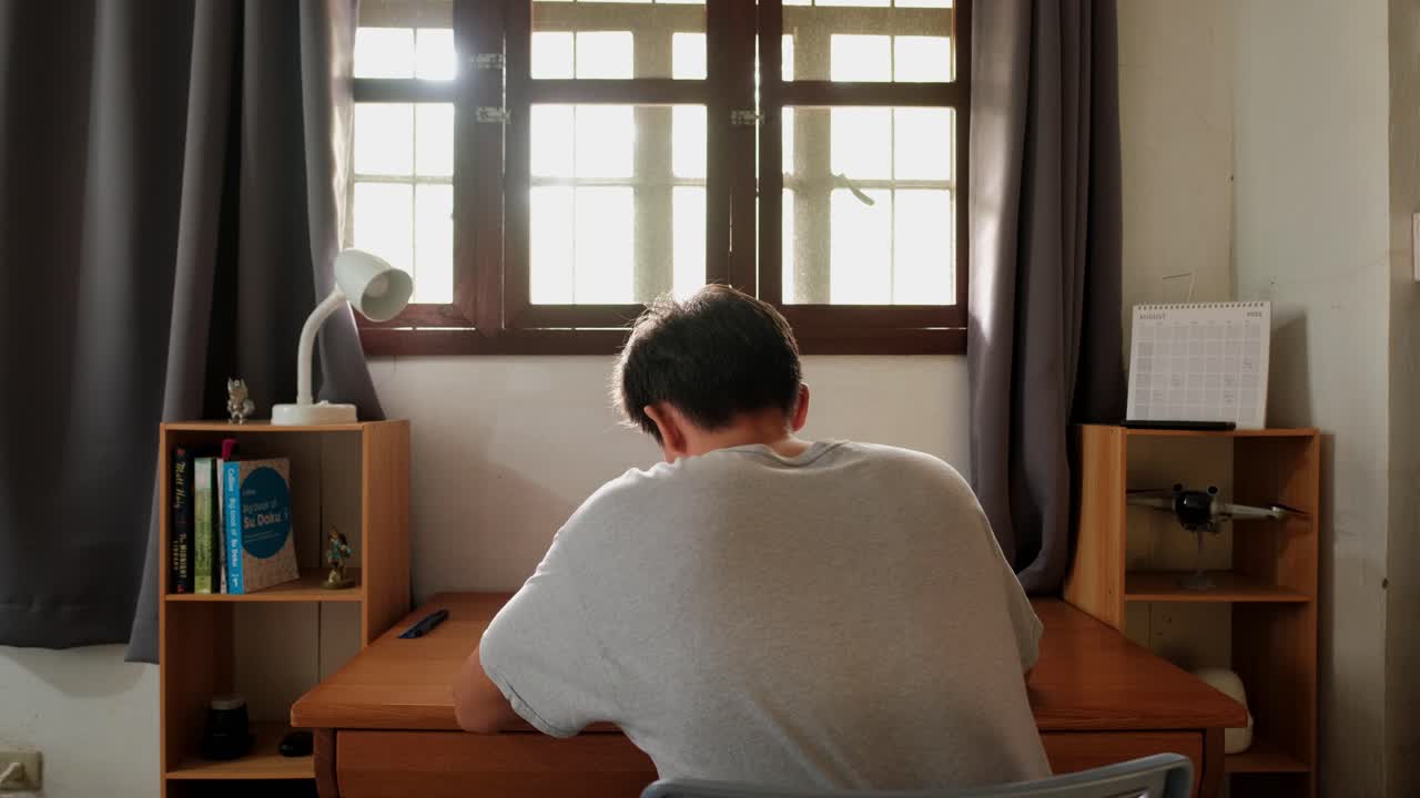 Static shot of young, male student writing in notebook and studying in front of idyllic sunlit window at bedroom desk during daytime