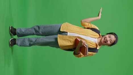 full body of asian woman student with a backpack and some books smiling and pointing to side while standing in the green screen background studio