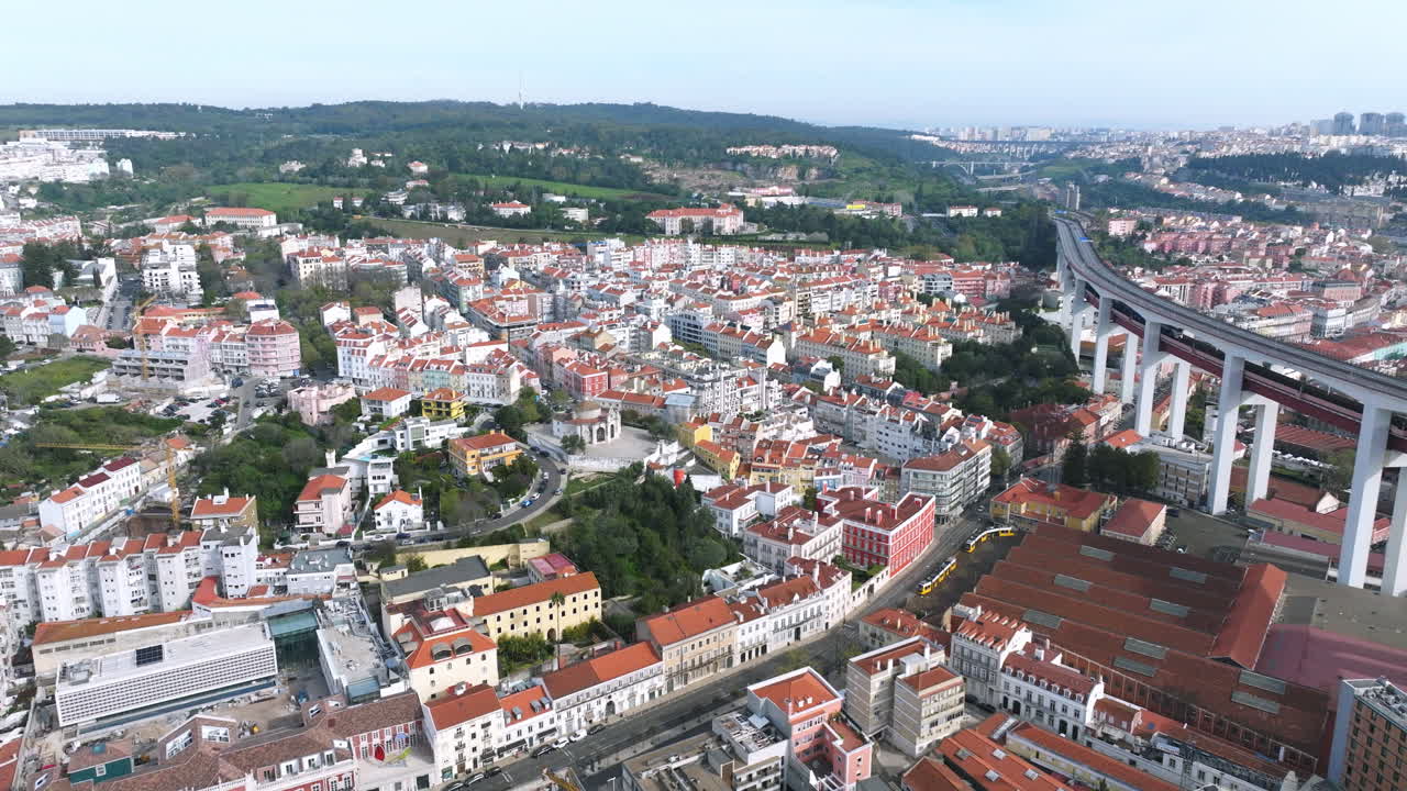 Aerial drone flight over the Lisbon cityscape on a clear morning, moving toward the Alcantara district and the green hills of Monsanto Park, with MACAM visible below