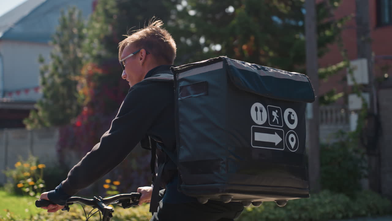 ciclista delle consegne bianco con zaino termico che pedala lungo una strada residenziale illuminata dal sole, espressione concentrata, borsa termica con icone, prospettiva ravvicinata, luce mattutina e ombre soffuse, ultimo miglio