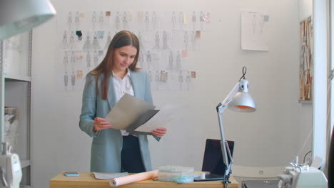 female fashion designer looking at drawings and sketches that are pinned to the wall behind her desk. studio is sunny. personal computer colorful fabrics sewing items are visible.