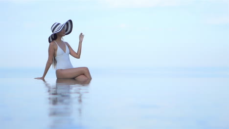 Eine-Attraktive-Junge-Frau-In-Einem-Badeanzug-Sitzt-Auf-Dem-Infinity-Rand-Eines-Swimmingpools-Und-Vermittelt-Die-Illusion,-Mitten-In-Der-Luft-Zu-Schweben