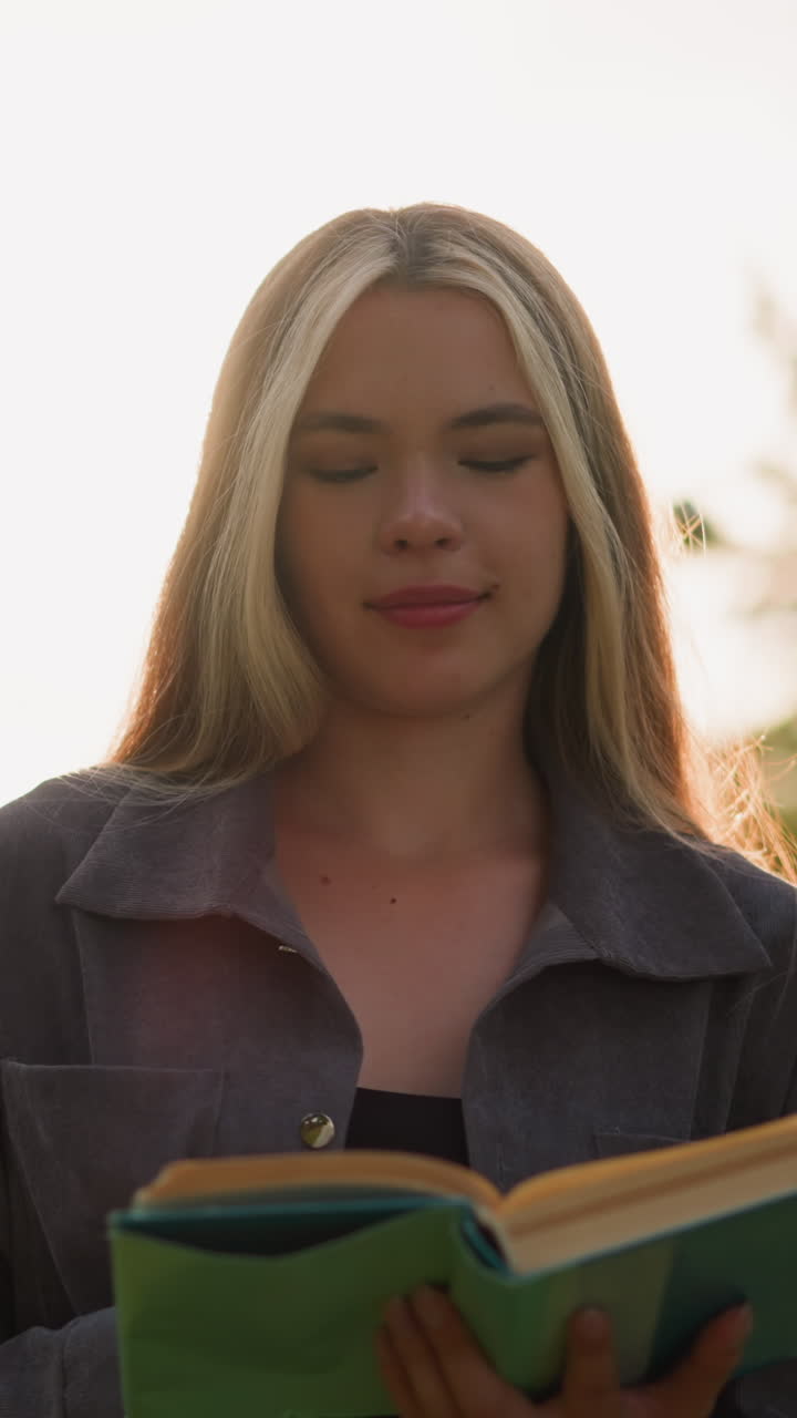 dama de ropa gris caminando por un campo de hierba mientras lee un libro mientras la luz del sol crea destellos brillantes a su alrededor, mirando pensativa con una cálida sonrisa, con un fondo iluminado por el sol borroso