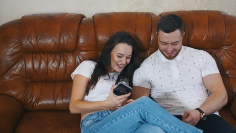 Cheerful friends playing on phones. Young couple sitting on a comfortable sofa and having fun together. Beautiful woman talking to her husband at home happily.