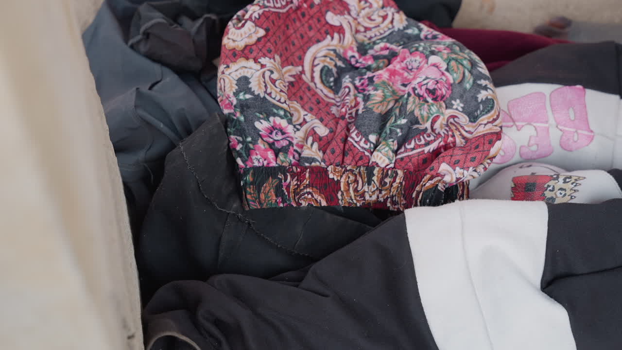 Scattered laundry on carpeted floor showing mixed garments, floral patterned fabric, towels and socks pushed beside couch, soft natural light, cozy interior, sense of everyday clutter and unfinished