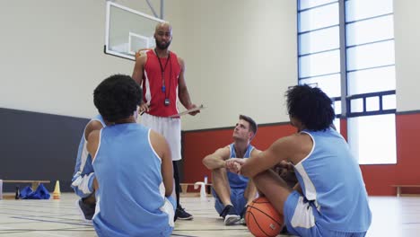 Entrenador-De-Baloncesto-Masculino-Diverso-Con-Portapapeles-Instruyendo-Al-Equipo-Sentado-En-La-Cancha,-En-Cámara-Lenta