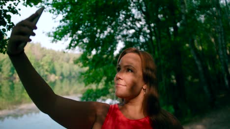 A-happy-woman-taking-selfie-photos-with-a-cellphone-with-a-view-of-the-lake-and-woodst-in-the-background