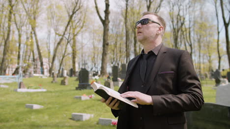 man in black suit and sunglasses reading a bible in a graveyard