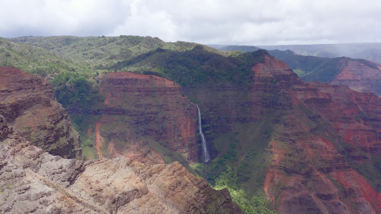 Smooth 4K drone footage of Waipoo waterfall in the background in the shade of a cloud overhead. In the foreground, dry, rocky peaks of the canyon wall in the bright sunshine