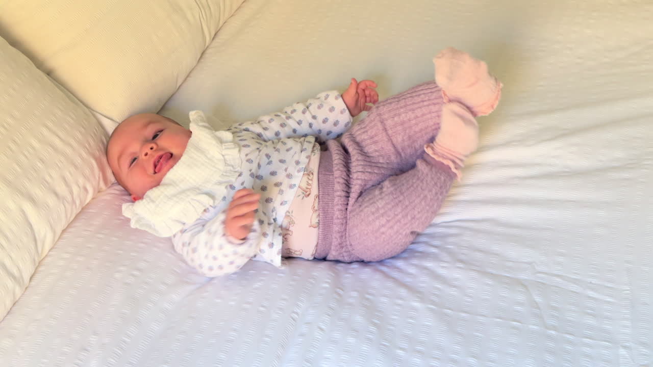 A sweet infant lies on a cozy bed, laughing while playfully moving hands and feet. Dressed in soft, pastel clothes, the little one enjoys a happy moment, creating a heartwarming and tender scene