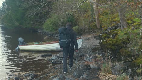 young man backpack island shoreline puts bag in lake boat wide shot