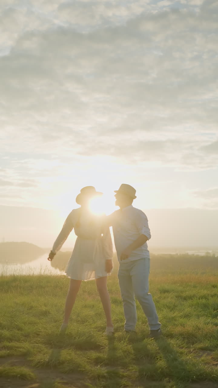 한 커플이 평온한 호수 근처의 잔디 언덕에서 한 에 춤을 추고, 황금빛을 즐기면서 안고 있습니다.  셔츠와 모자를 입은 남자, 흐르는  드레스를 입은 여자.