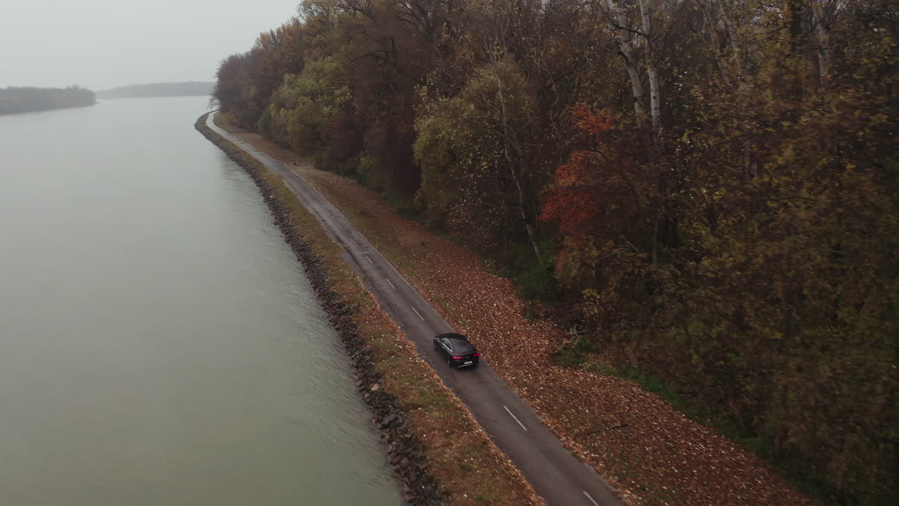 Inseguimento aereo con drone di un'auto di lusso nera che guida sotto la pioggia - Sponda del fiume in campagna - Cielo coperto autunnale