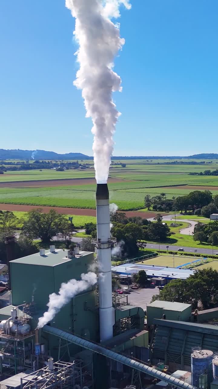 Un filmato con drone mostra un'alta ciminiera industriale che rilascia vapore bianco sopra un impianto di lavorazione rurale.