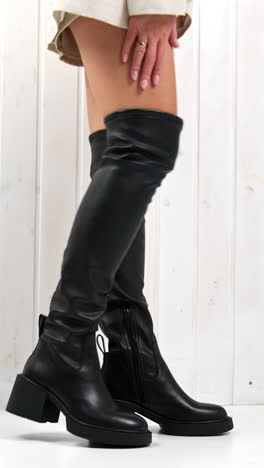 woman touching the high black boot on her leg. lady demonstrating the fashionable footwear in studio. white backdrop. low angle view. vertical video