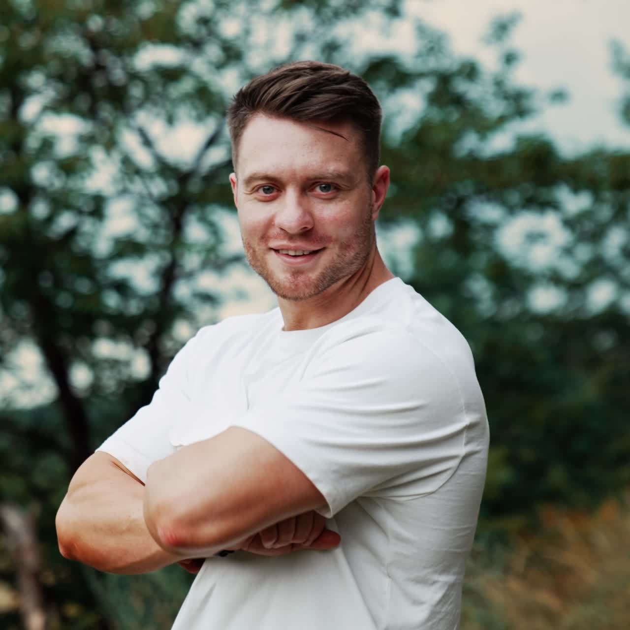 Confident mid-aged Caucasian man wearing white shirt. Smiling muscular man folds hands on the chest looks at camera. Blurred nature backdrop