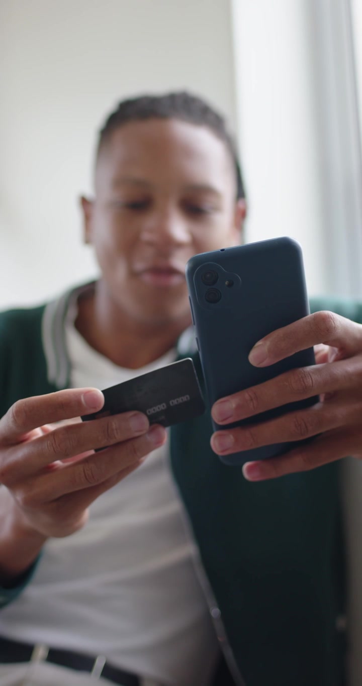 Vertical video: African man lifting black card resting on thigh on couch, scanning it with phone