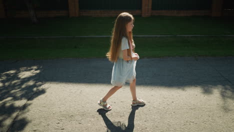 sober young girl with long hair walks slowly while holding skirt in one hand, moving along shaded street with green fence and brick pillars in background