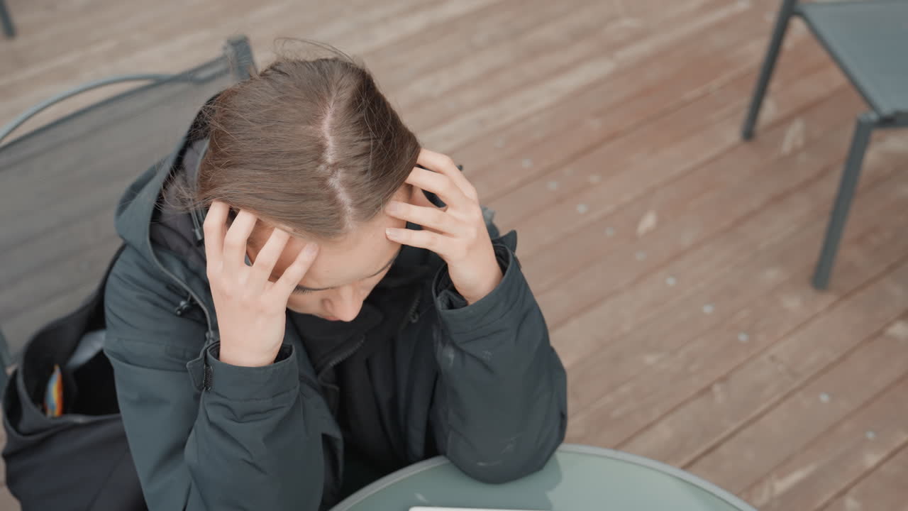 Donna caucasica seduta all'aperto in un bar, che si sistema le cuffie e poi si prende la testa tra le mani per la frustrazione. Indossa un cappotto invernale su una terrazza in legno, con uno smartphone su un tavolo rotondo, sedie vuote sullo sfondo, cielo nuvoloso e un'espressione tesa.
