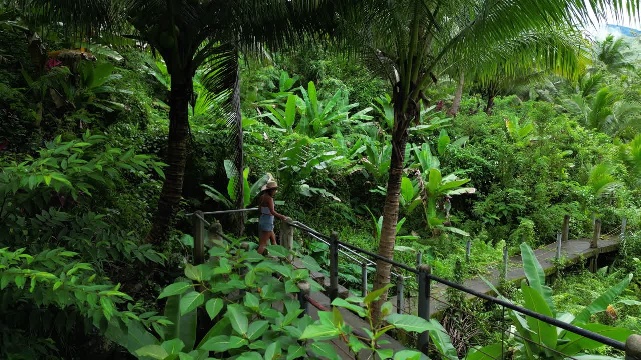 Back-tracking aerial of woman walking elevated metal bridge through dense tropical foliage, gently brushing railings as she moves deeper into the forest in Tanay Rizal, Philippines