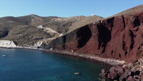 orbita aérea playa roja llena de turistas y acantilado empinado, santorini, grecia