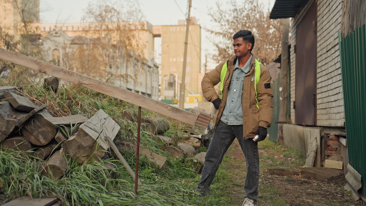 Uomo che ispeziona il legname prima della riparazione in un cortile urbano, giacca ad alta visibilità e guanti, appoggiato a una tavola recuperata mentre valuta l'allineamento della trave, pianificazione e valutazione ponderate, calmo e fiducioso.