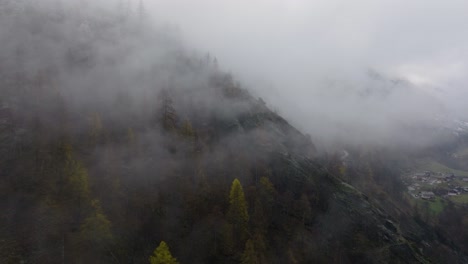Mysterious-aerial-forwards-rotating-in-Swiss-Alps-on-cloudy-moody-grey-winter-afternoon-over-lush-green-pine-forests-and-beautiful-snowy-trees-on-mountains-sideways-flying-through-town-valley
