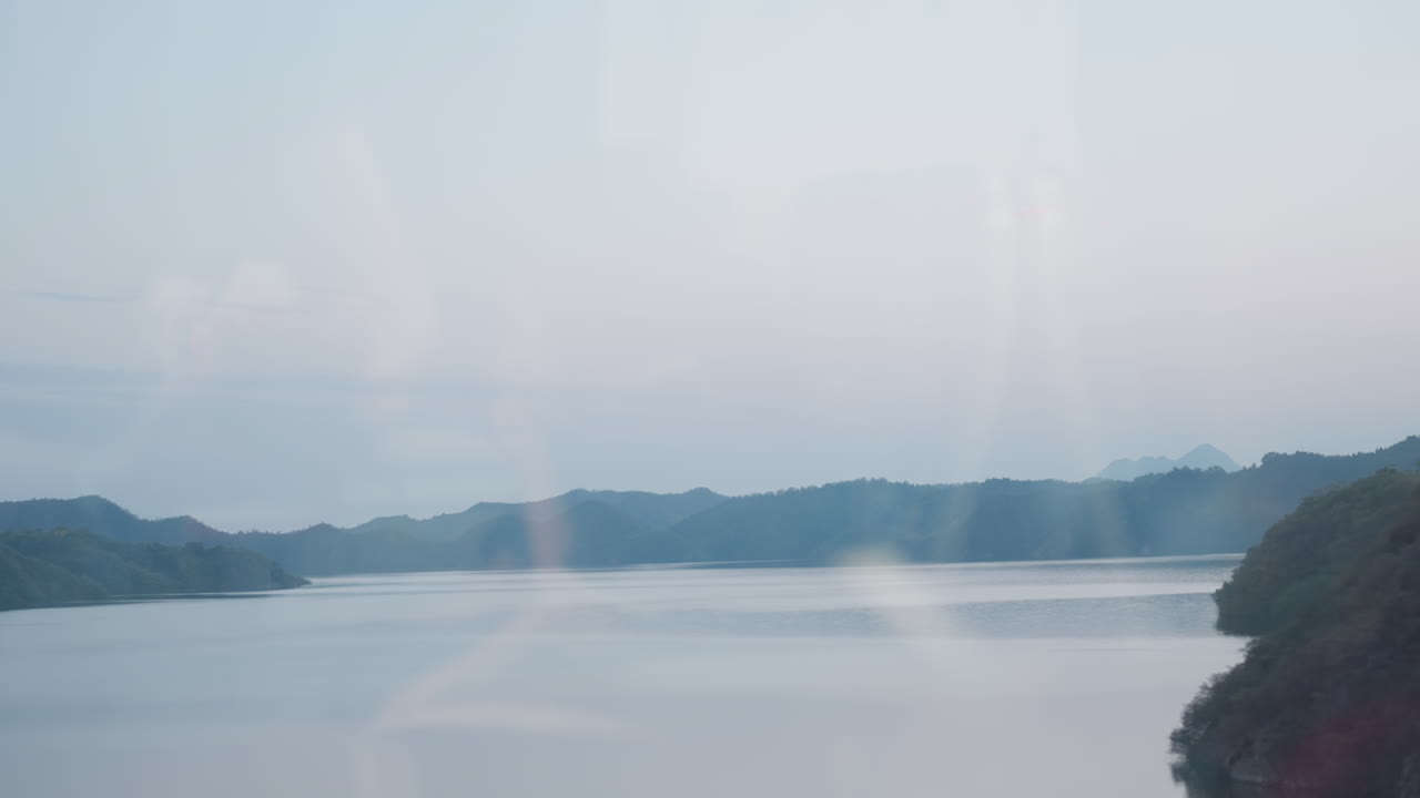 電車の窓から見える湖の景色、スマートフォンを使う乗客の姿が映り込み、座席や吊り革はぼやけている。遠くの島々と穏やかな水面、パステルカラーの夕暮れ空が広がり、モーションブラーが物思いにふける雰囲気を伝えている。