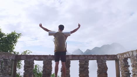 asian hiker male standing on a veranda cliff enjoys seeing beautiful view of top foggy mountain and raising his hands. a man from the tent view of the mountain