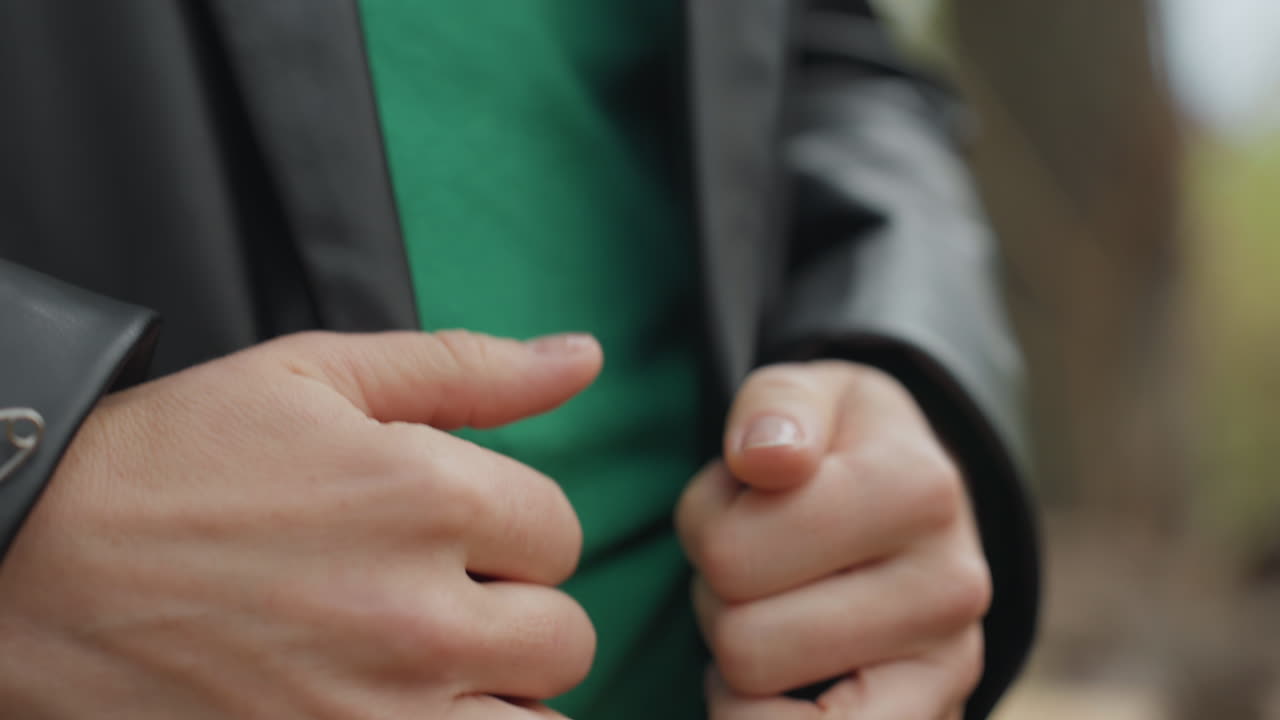 Primo piano di donna caucasica che sistema una giacca di pelle sopra un maglione verde in un ambiente naturale all'aperto, dita che si agitano, pollici che lisciano il polsino, movimento della cerniera, mani che stabilizzano il busto, sfondo bokeh autunnale